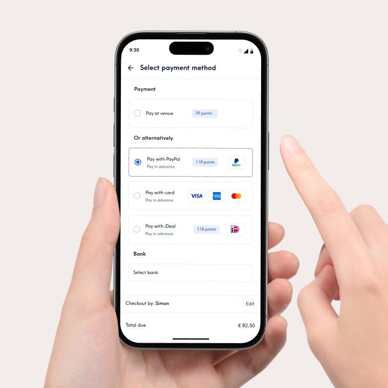 Close-up of a smartphone showing the payment interface of the Treatwell app for selecting a payment method. Options include paying at the venue or paying in advance with PayPal, card or iDeal. A hand holds the phone and a finger points at the screen.