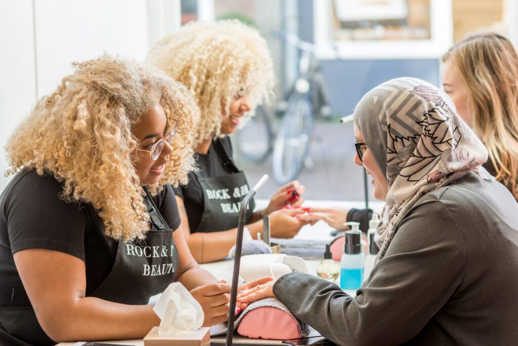 Two manicurists with blonde curly hair wearing black aprons that read "Rock & Roll Beauty" are giving manicures to clients at a long table. One client is wearing a grey hijab.