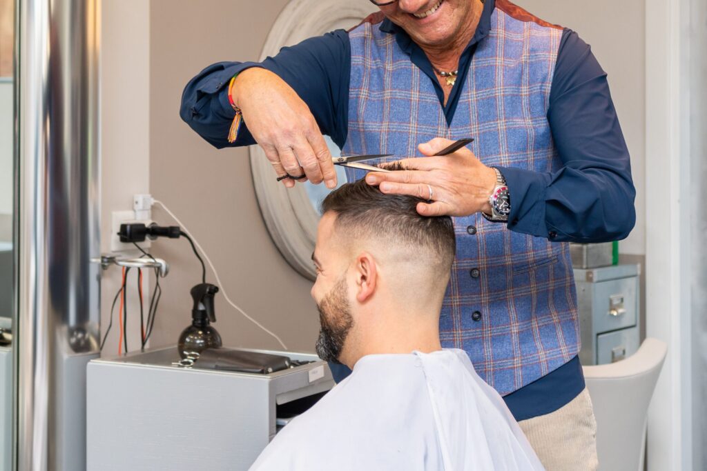 A male hairdresser wearing a blue checkered waistcoat cuts a client's hair with scissors and a comb in a salon. The client has short hair and a beard, and is wearing a white hairdressing cape.
