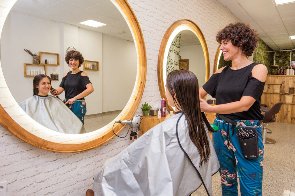A smiling female hairdresser with short curly hair brushes a client's long damp hair in front of a large round illuminated mirror. The scene is reflected in the mirror, showing the salon interior.