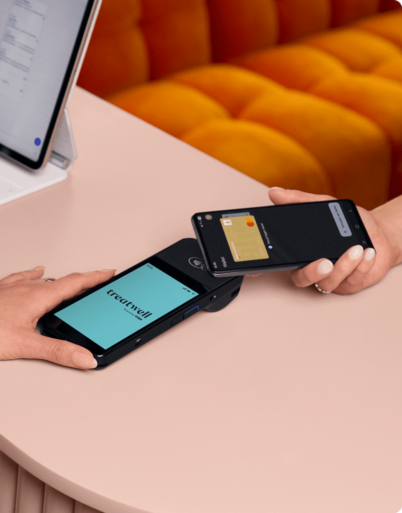 A high-angle close-up of a contactless payment being made. One person holds a Treatwell POS card machine displaying the brand logo, while another person taps a smartphone over it to pay. The background features a bright orange velvet sofa.