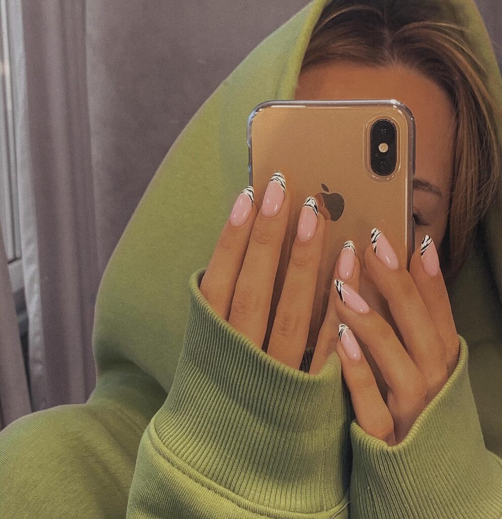 Person in green hoodie holding phone, showing manicure with pink nails and black-and-white striped French tips. Credits @sansilt
