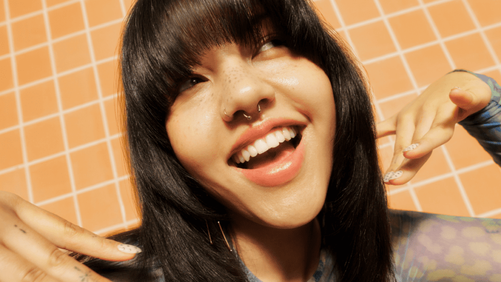 Smiling person with straight bangs and septum piercing, showcasing trendy nails against a tiled background.