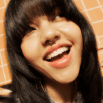 Smiling person with straight bangs and septum piercing, showcasing trendy nails against a tiled background.