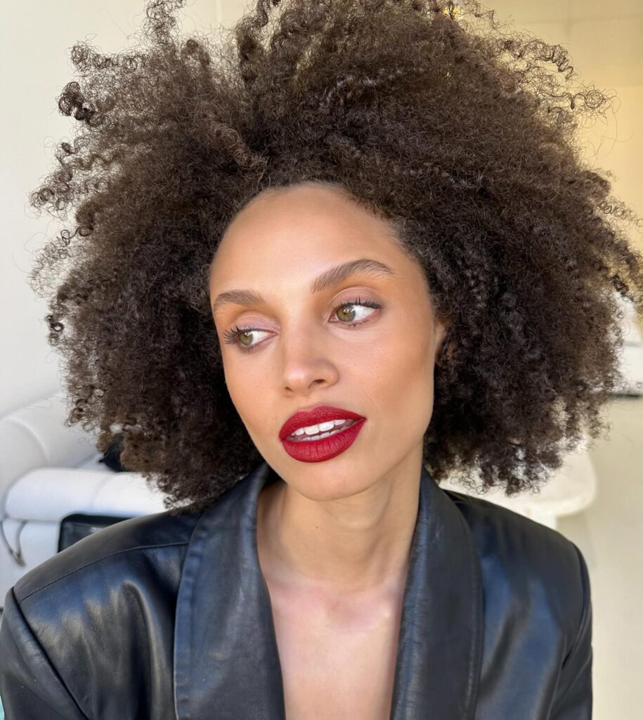 Woman with natural curls and bold red lipstick wearing a black leather jacket, embodying spring 2026 beauty trends. Credits: @tannia_t_mua