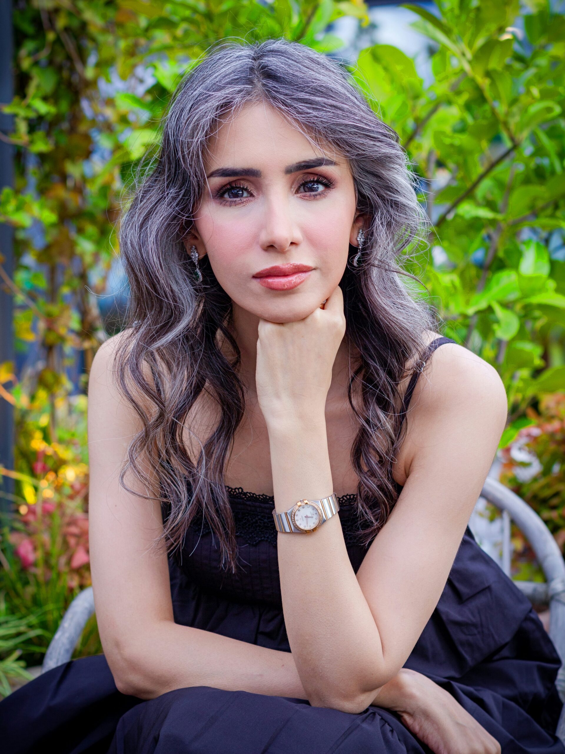 Woman with long wavy dark hair and blended grey sits on a chair in a garden and poses with her hand on her chin