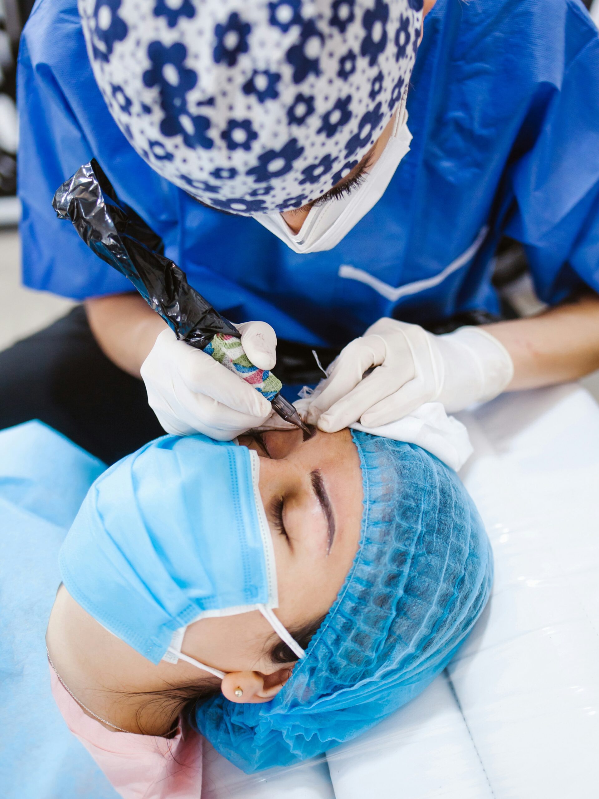A close-up of a woman receiving microblading treatment from a technician, she lies on a treatment bed and wears a mask