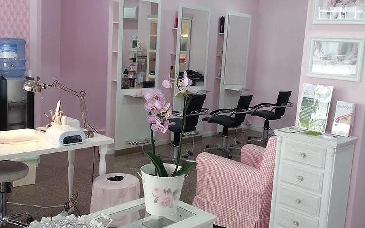 The interior of a bright, pink-themed hair and nail salon. On the left, a white manicure station is equipped with a desk lamp and nail curing device. In the centre, an orchid plant in a white pot sits on a coffee table next to a pink-and-white gingham armchair. The background features several hair styling stations with tall mirrors and black salon chairs against light pink walls.