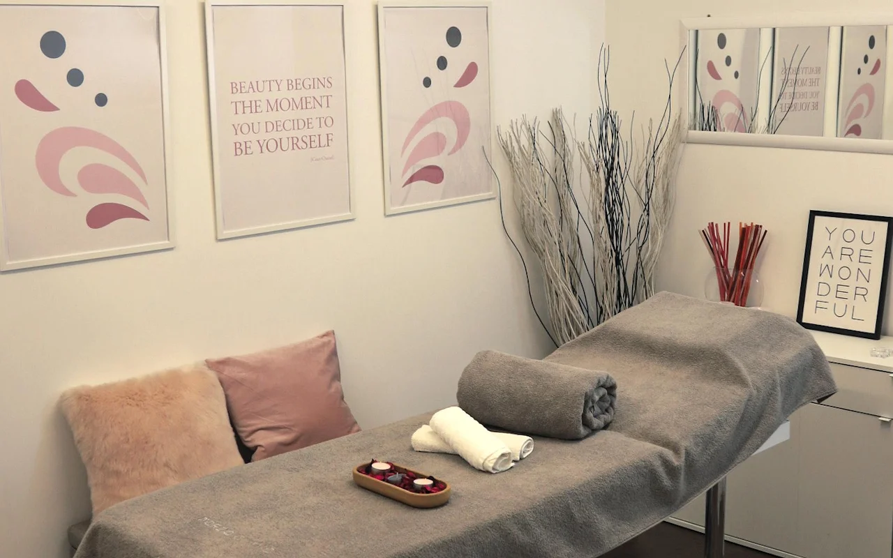 A bright treatment room with a massage table draped in a grey towel. The wall is decorated with pastel-toned abstract art and motivational quotes, while decorative white branches and candles create a calm atmosphere.