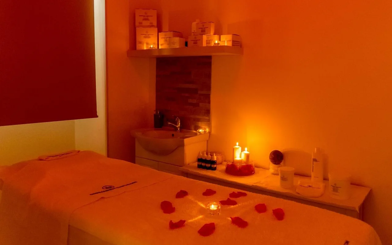 A massage room with warm, moody orange lighting and flickering candles. Red rose petals are arranged in a circular pattern around a central candle on the treatment table, with beauty products neatly organised on a shelf behind.