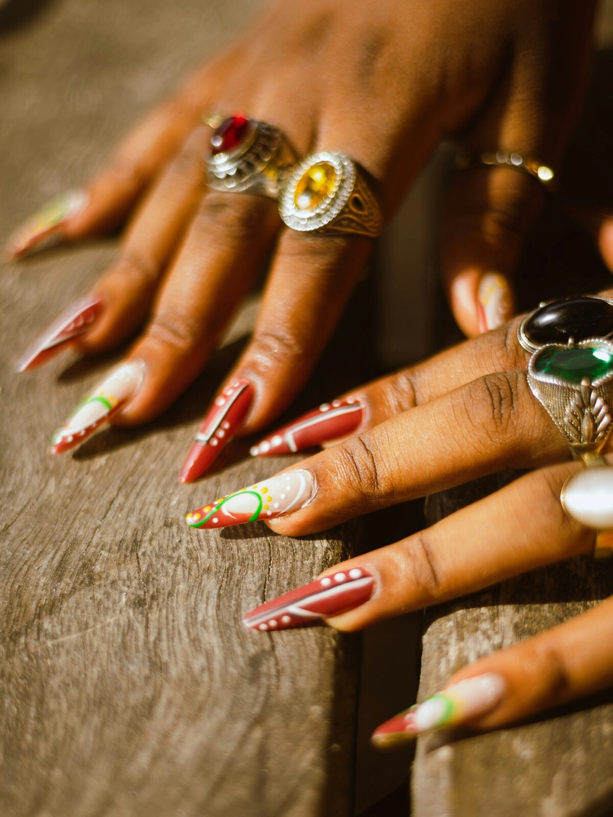 Close-up of a woman’s hands with stiletto-shaped acrylic nails in red and white decorated with green and yellow dots and swirls