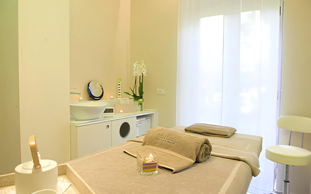 A calm treatment room at Alice Beauty Clinique with a bed dressed in soft taupe linens, white orchids on the counter, candles by the basin, and sheer curtains filtering natural light through a large window.