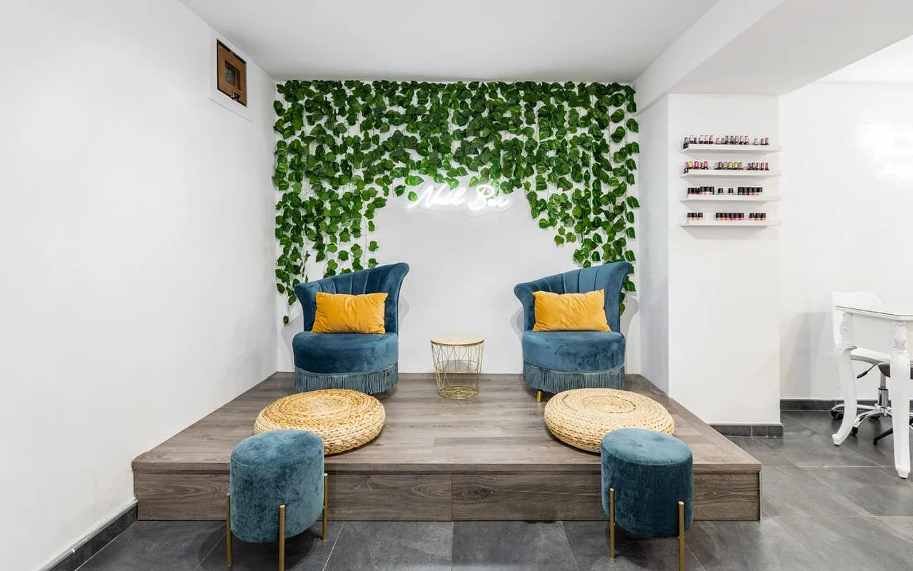 A nail bar corner with a cascading green foliage wall and a small neon sign, two teal velvet armchairs with mustard cushions on a raised wooden platform, woven footstool pouffes, and shelves of colourful nail polish on the right.
