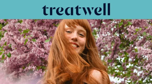 A woman with long copper-red hair and a fringe smiling in the sunshine, with pink blossom trees in the background. The Treatwell logo sits on a teal banner above.