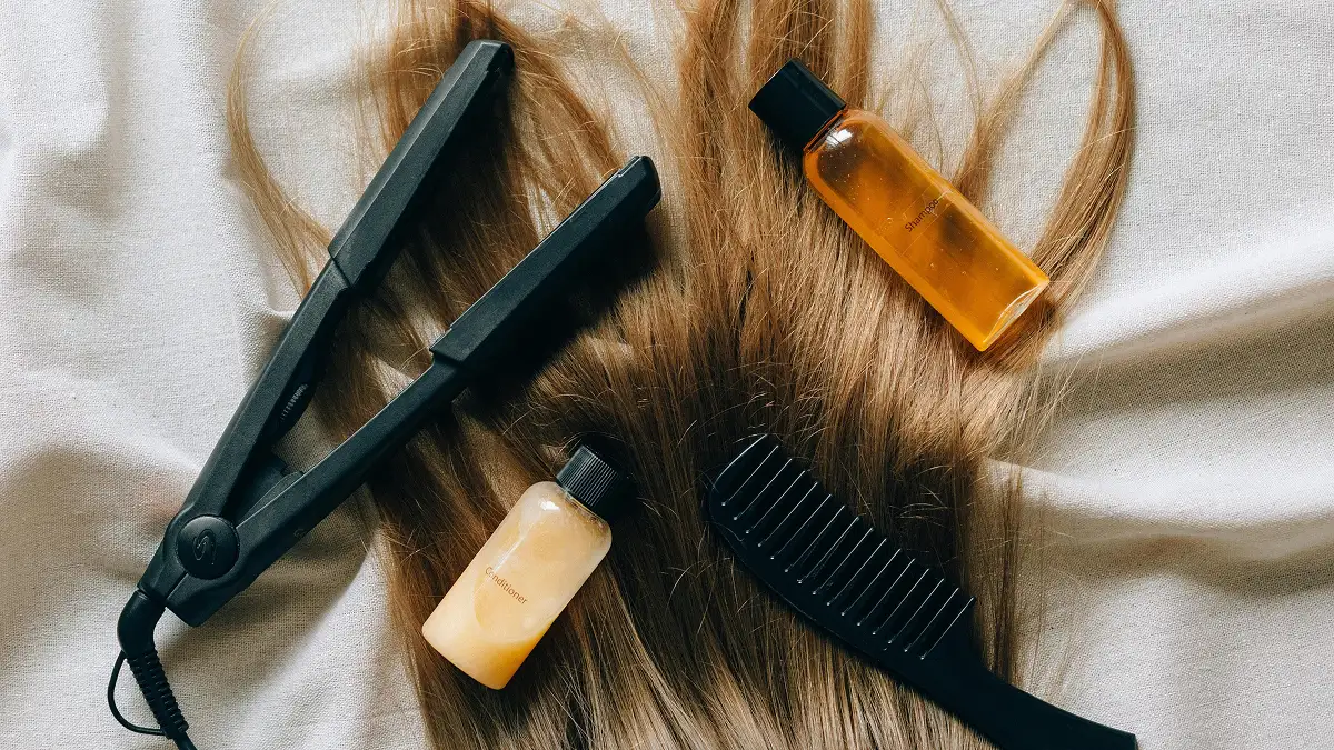 Flat lay of hair care products arranged on top of someone’s long straight hair, including shampoo, conditioner, straighteners and a wide tooth comb