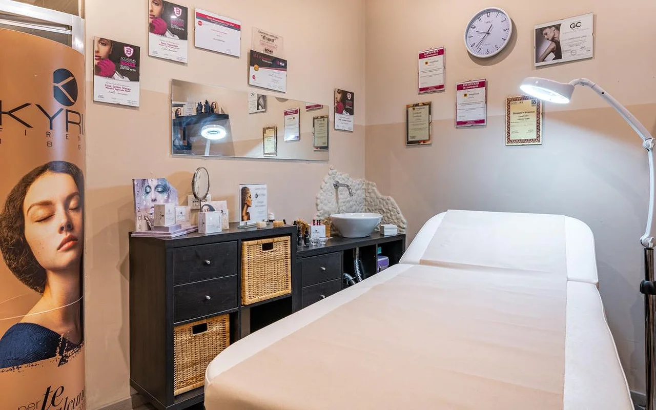 A treatment room at Oltre la bellezza with a white treatment bed, adjustable magnifying lamp, and walls lined with professional certificates and diplomas. Dark wicker storage units sit beneath a mirror, with skincare products arranged on the counter.