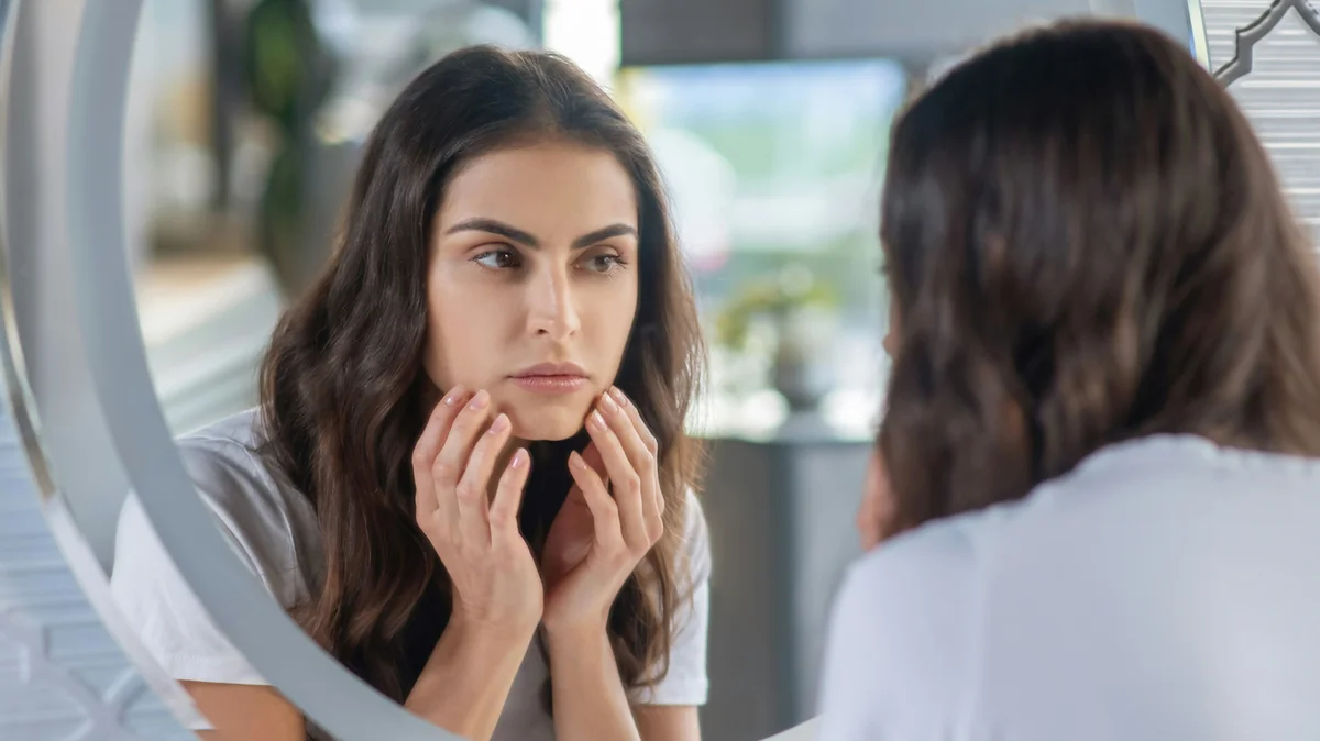 A woman with long dark hair and neat, well-defined brows looks at herself in the mirror, holding her hands gently to her face