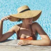 A woman in a sunny outside swimming pool leans on the edge with her arms, she wears a burnt orange bikini and a wide-brimmed straw sun hat