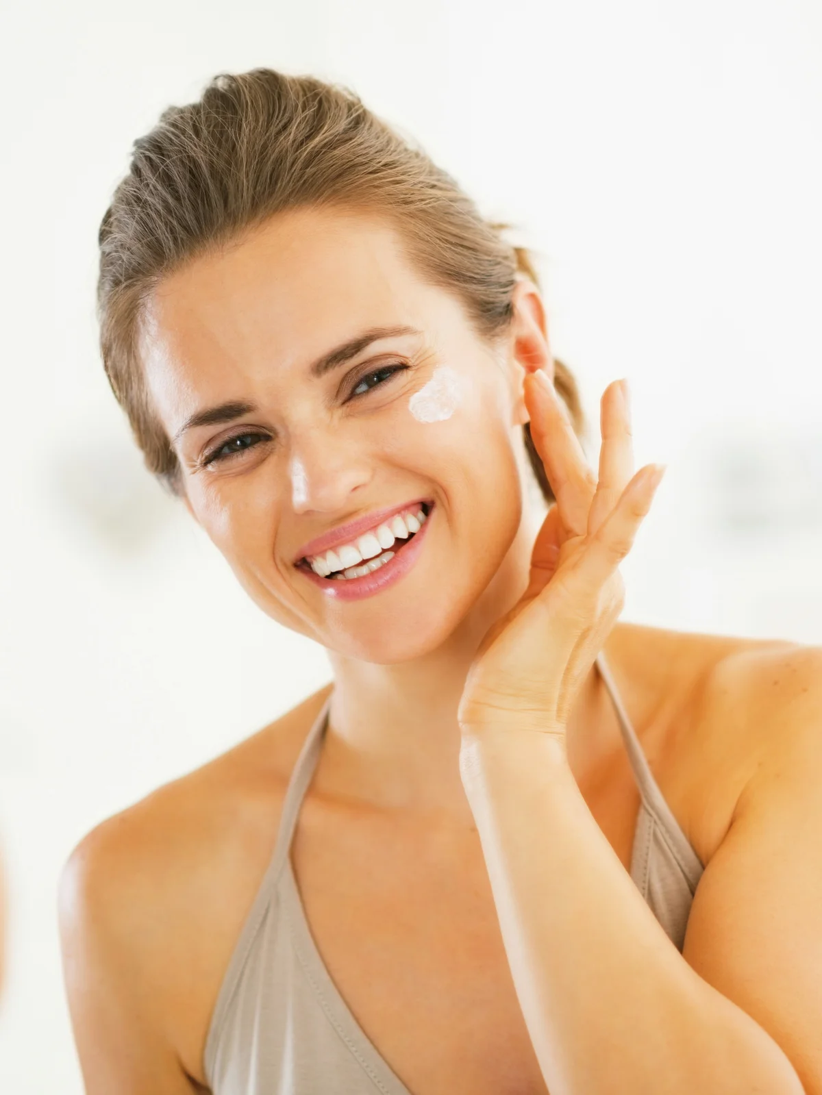 Close-up of a smiling woman with her hair tied back applying SPF to her face