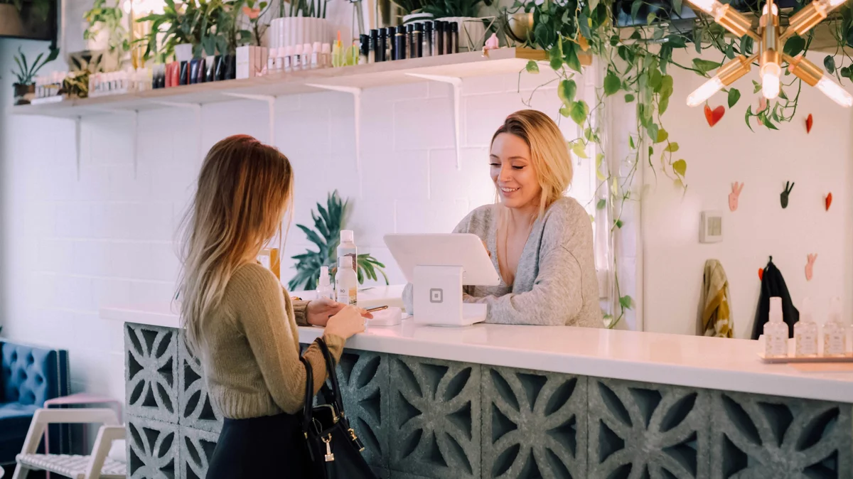 A woman stands at a salon desk where a receptionist books her next treatment, the salon is clean with a white desk, neat shelves of products and plants and flowers decorating the space