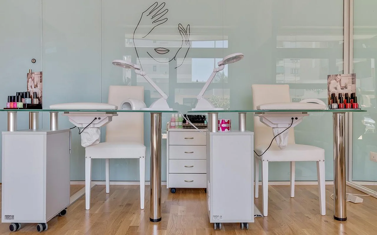 A nail studio at Perfetta Urban Beauty, with two glass-topped manicure tables and white chairs, task lamps, colourful polish displays, and a mint-green glass wall bearing a line drawing of a face and hands.