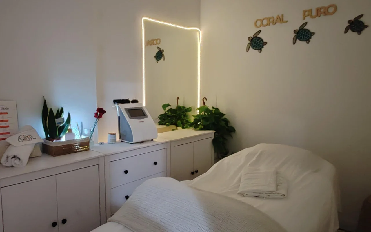 A small, calm treatment room with white cabinets and a bed dressed in white towels. An illuminated mirror and a treatment machine sit on the counter among green plants, below gold 'Coral Puro' lettering and decorative turtle wall art.