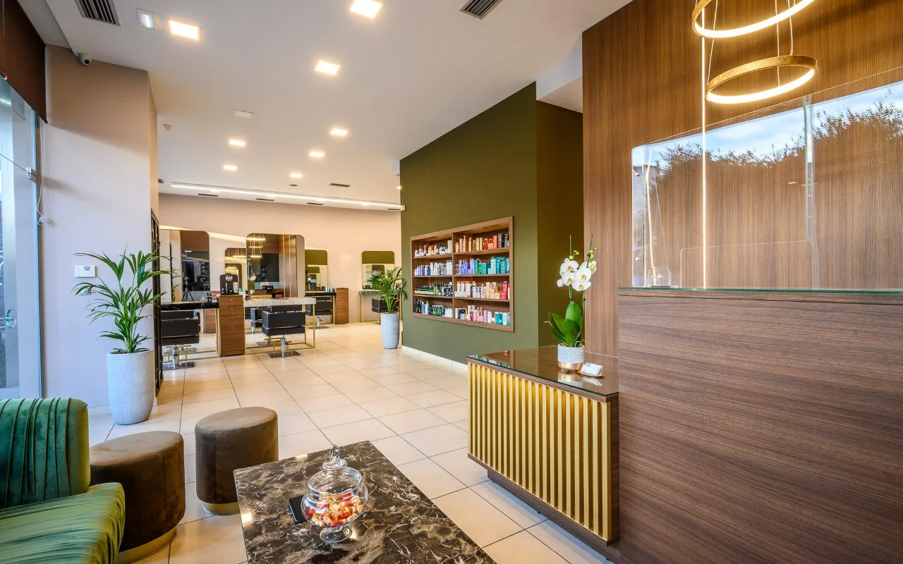 An upmarket hair salon with warm wood panelling, an olive-green feature wall of product shelves, styling stations with large mirrors, a gold-slatted reception desk and green velvet seating beneath circular gold pendant lights.