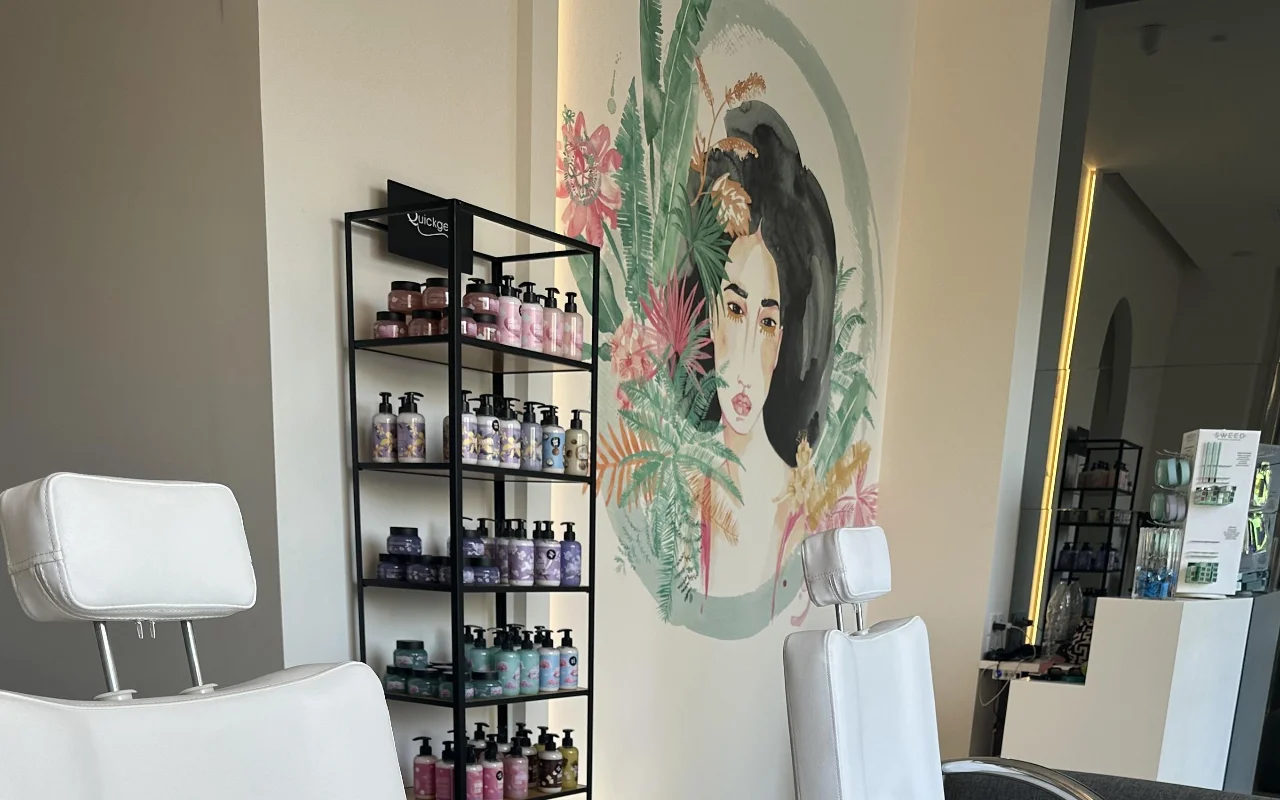 A studio corner with two white reclining treatment chairs, a black shelving stand of products, a backlit mirror, and a floral wall mural of a woman's face framed by tropical foliage.