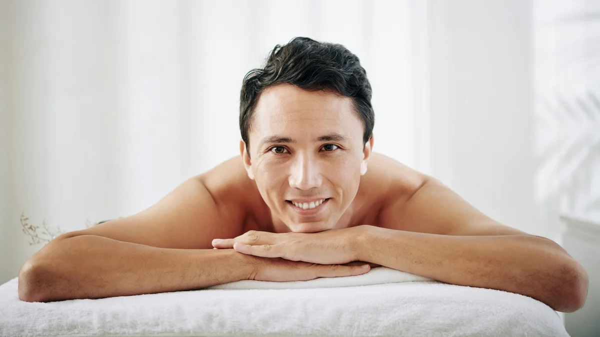 A man lays on his front on a treatment bed at a beauty salon, he rests his chin on his hands and smiles