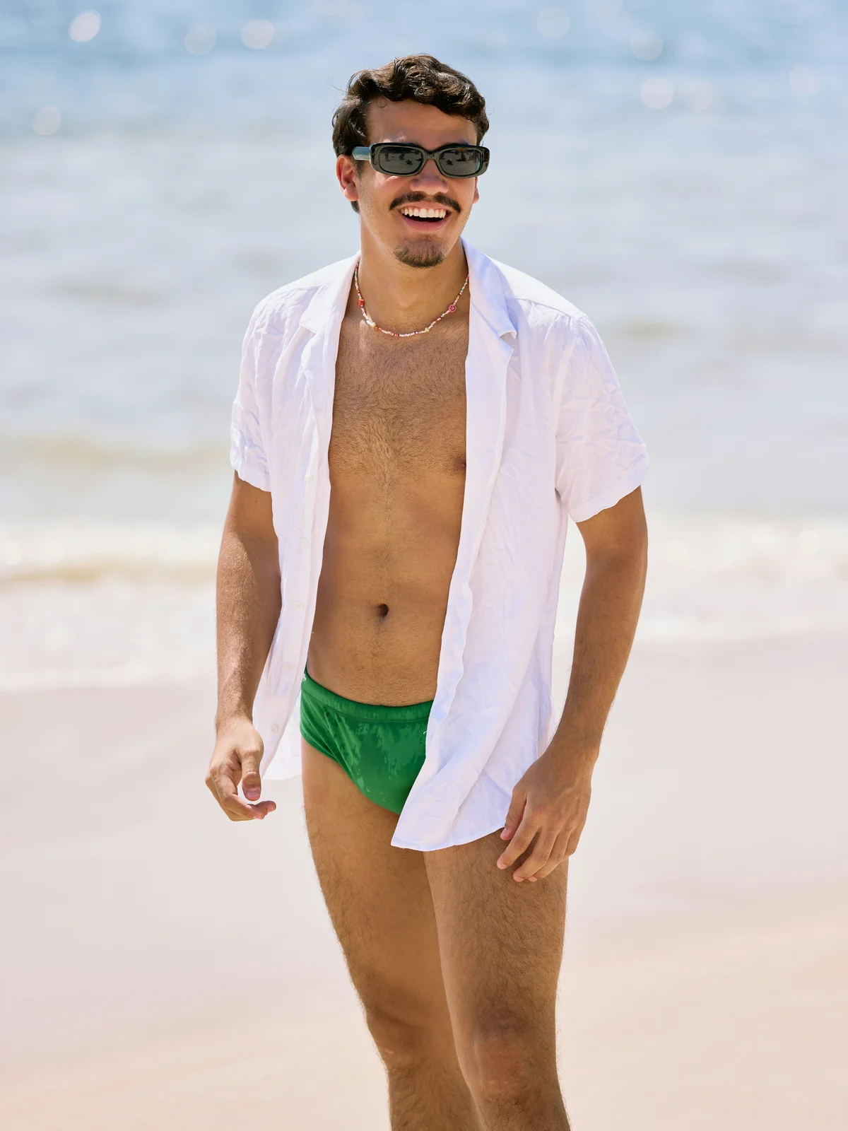 A smiling man stands on a beach on a sunny day wearing green swimming pants, an unbuttoned white shirt and black sunglasses
