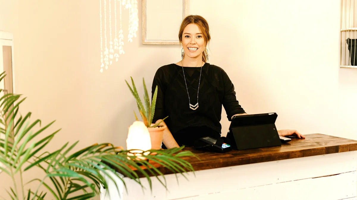 A smiling woman stands behind a reception desk at a beauty salon
