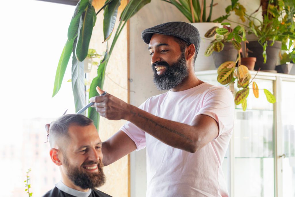 Een glimlachende kapper met een donkere baard en een grijze platte pet gebruikt een schaar en een kam bij het haar van een glimlachende klant in een licht salon vol groene kamerplanten.