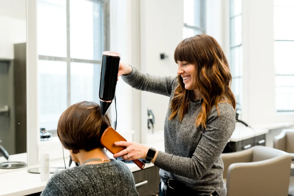 Een lachende kapster föhnt en borstelt het haar van een klant in een lichte, moderne salon.