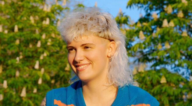 Een jonge vrouw met een krullend witblond mullet-kapsel zittend buiten op een zonnige dag.