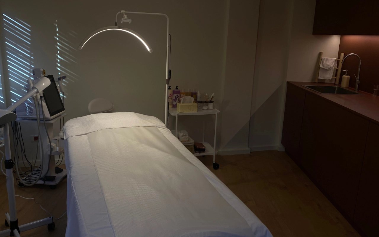 Een gerichte behandelkamer met een wit bed onder een boog-LED-lamp, professionele aesthetics-apparatuur met een digitaal scherm, huidverzorgingsproducten op een trolley en donkerhouten kasten met een wastafel langs de achtermuur.