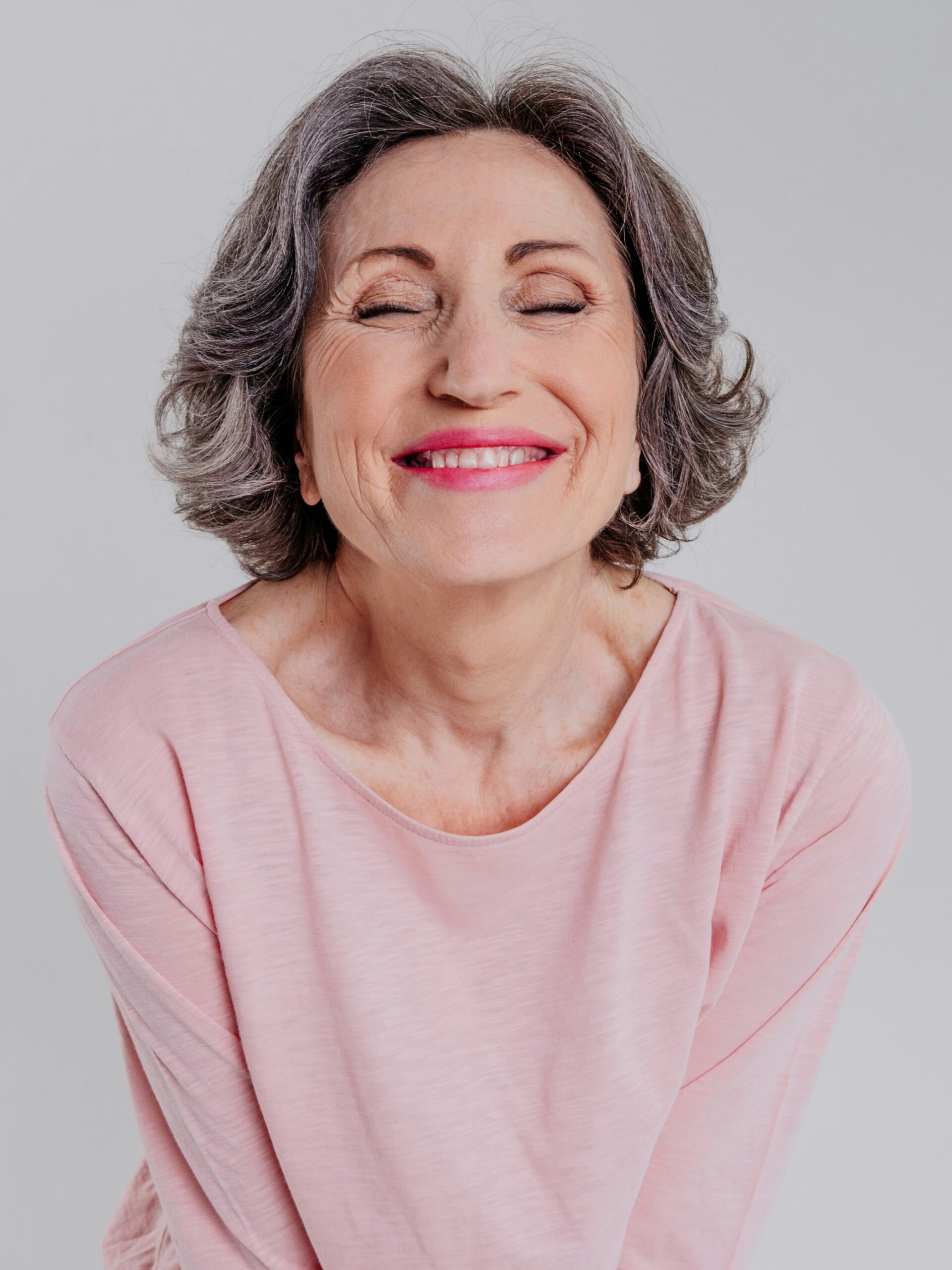 Portret van een vrouw met kort golvend salt-and-pepper haar in een los roze top, lachend met gesloten ogen