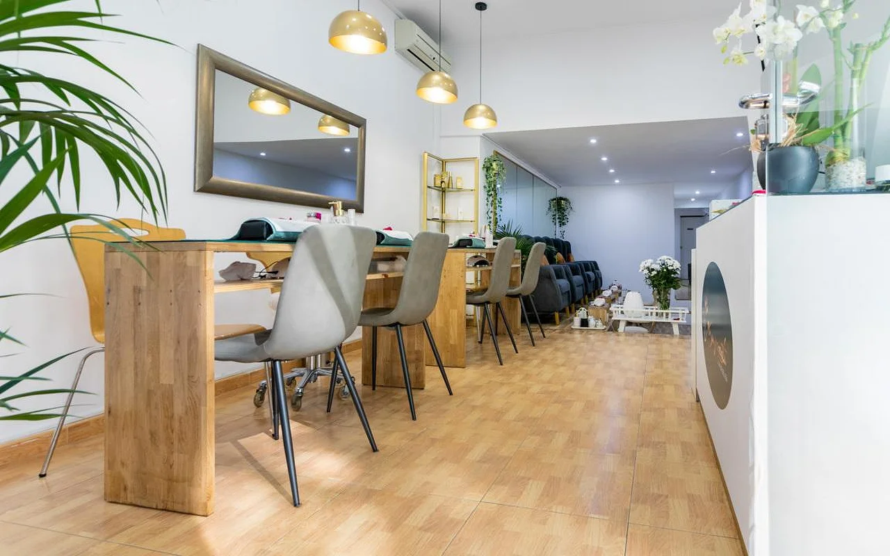 Een lichte nagelsalon met lange houten manicuretafels en grijze moderne stoelen. Goudkleurige koepelhangers hangen boven de werkplekken, en een grote spiegel met een donker frame hangt aan de witte muur.