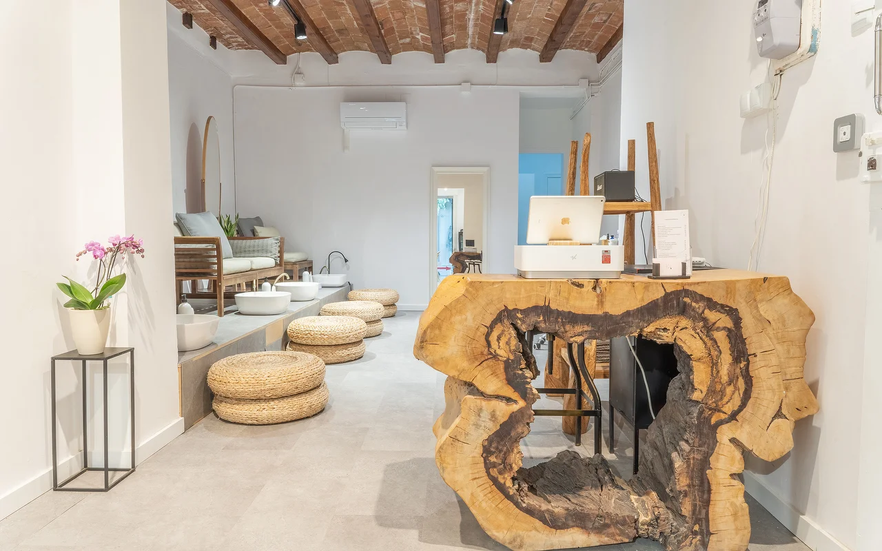 Een minimalistische wellnessruimte met een receptiebalie gemaakt van een grote, rauwe boomstamdoorsnede. Op de achtergrond staat een pedicurestation met geweven vloerkussens en witte bassins onder een rustiek balkenplafond.