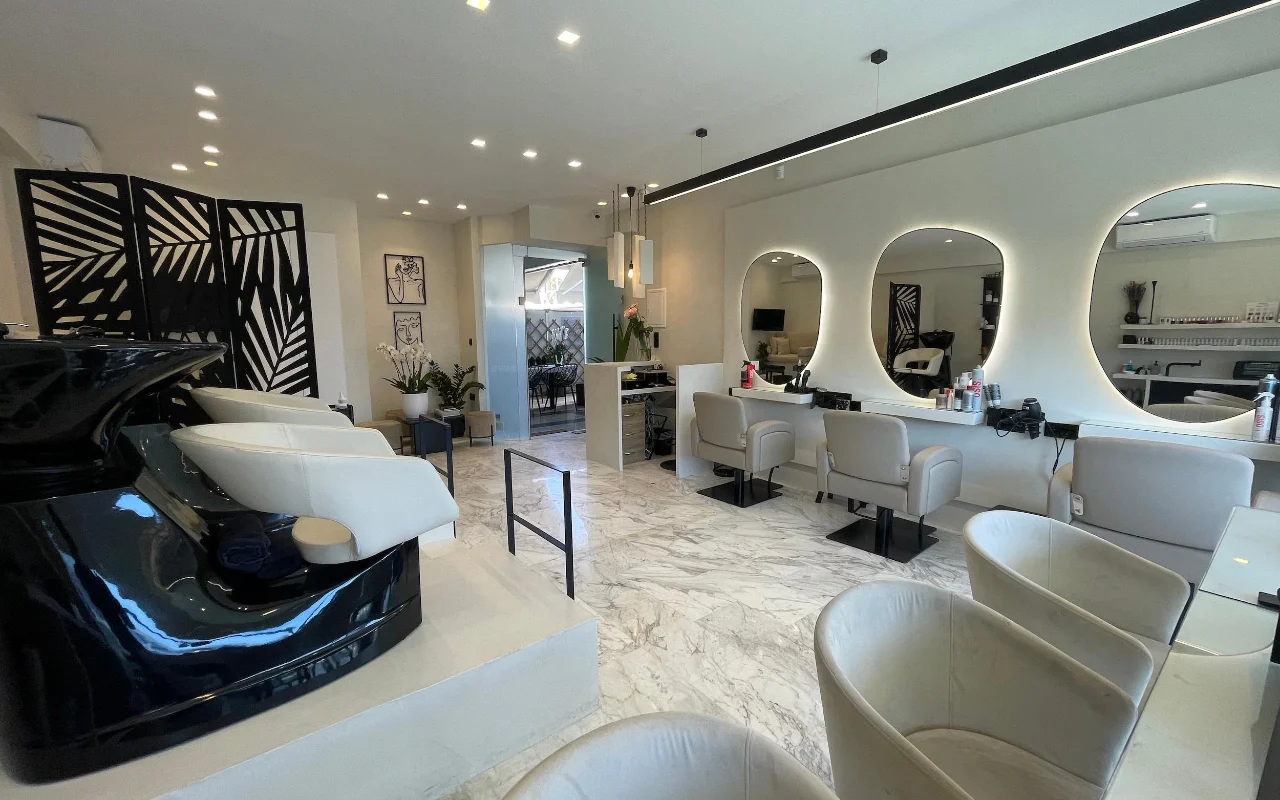 Een strakke haarsalon met witte marmeren vloeren, crèmekleurige leren stylingstoelen onder LED-omrande ovale spiegels, een glanzend zwart wasstation en een geometrische palmbladkamerscherm.