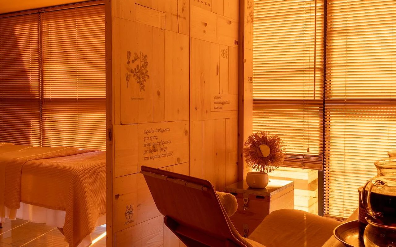 Een warme, amberkleurig verlichte spa-behandelkamer met natuurlijke houten wandbekleding, botanische illustraties en Griekse tekst in de muur gegraveerd, een behandelbed met een wafelstructuur-deken, en het APIVITA-logo in de hoek.