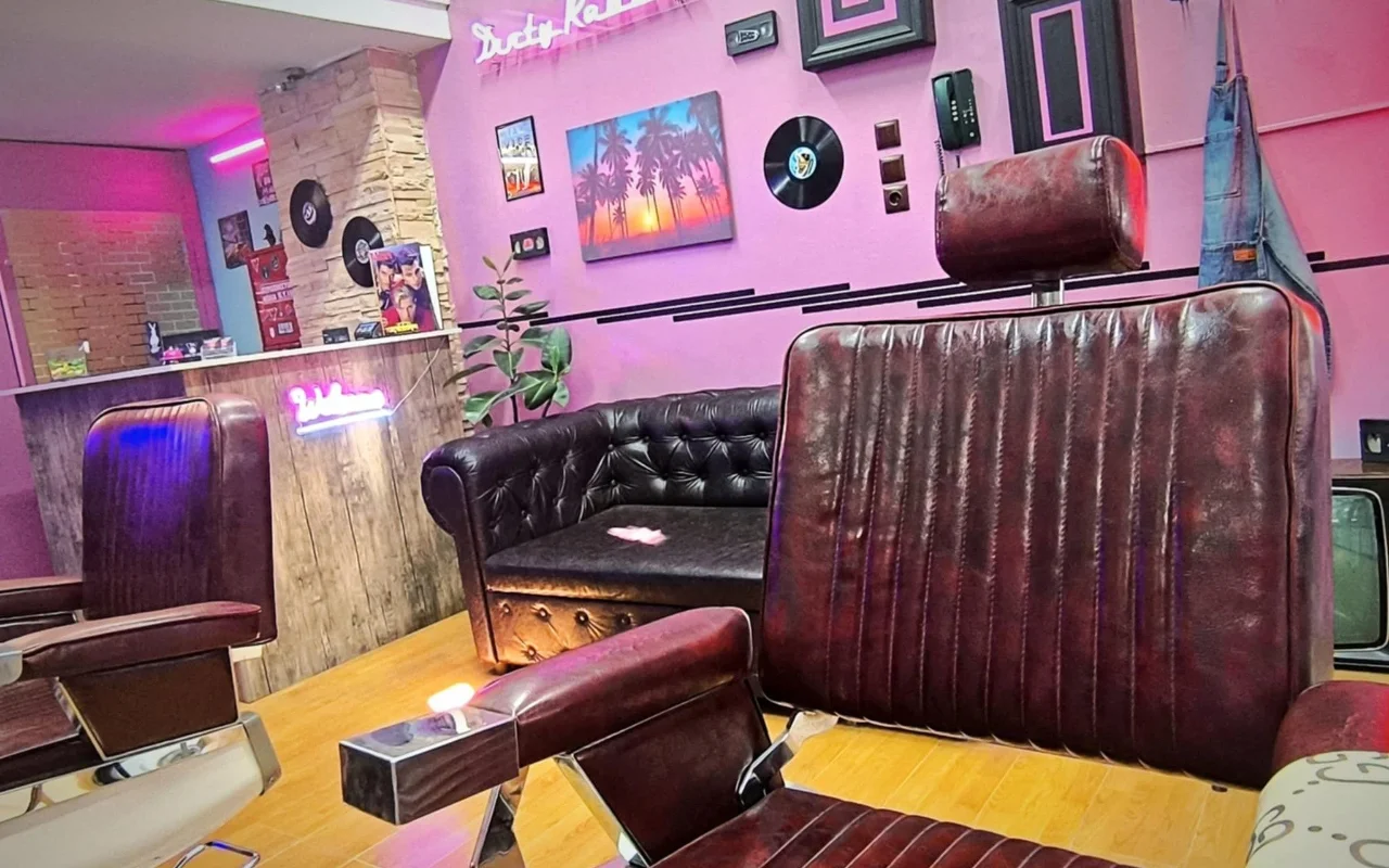 Een retro-stijl barbershop met roze en paarse muren, neonbelettering, vinylplaten als wanddecoratie, een schilderij van palmbomen bij zonsondergang, donkere leren kappersstoelen en een Chesterfield-bank op een warme houten vloer.
