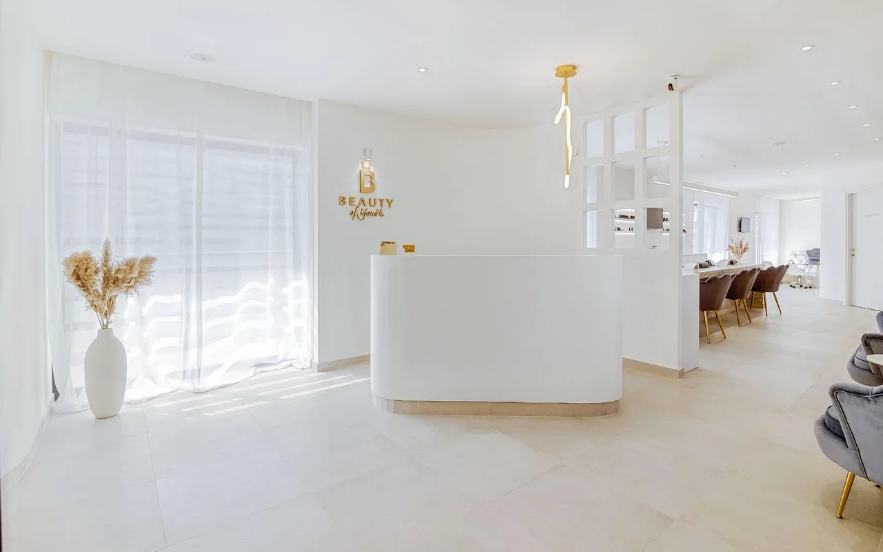 Een lichte, volledig witte ontvangstruimte met een gebogen balie, een gouden Beauty of Youth-logo op de muur, een sculpturale hanglamp, vitrage die natuurlijk licht filtert, en bruine fluwelen behandelstoelen zichtbaar op de achtergrond.