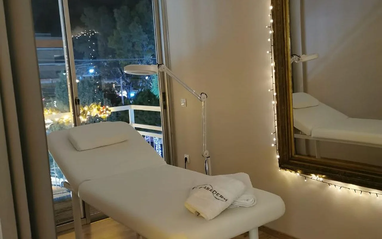 Een zacht verlichte behandelkamer met een wit bed, een handdoek met CasaDerm-logo, een vergrootlamp, lichtjes rond een gouden spiegel, en een balkondeur met uitzicht op het Glyfada-avondstraatbeeld.