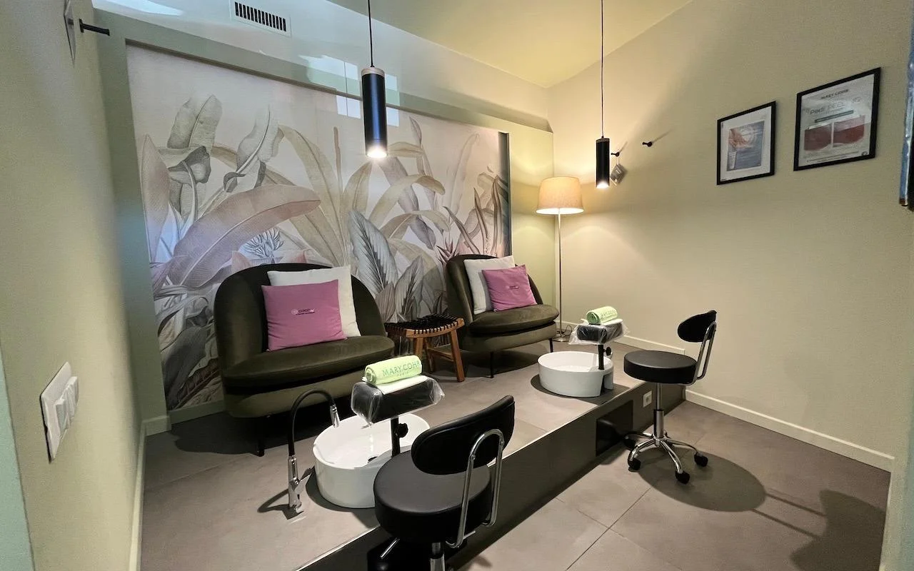 Een stijlvolle pedicurestation met twee diepgroene fluwelen fauteuils en roze kussens. Witte voetbaden zijn ingebouwd in een grijs verhoogd platform, tegen een muurschildering met grote tropische bladeren op de achterwand.