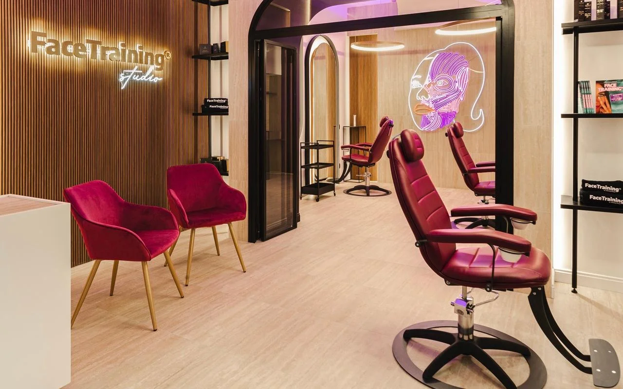 Een minimalistische studio met een houten latjes-wand en een verlicht logo. De behandelruimte heeft professionele bordeauxrode leren stoelen en een neon-lichtinstallatie met een gestileerd gezichtsspierdiagram op een glazen scheidingswand.