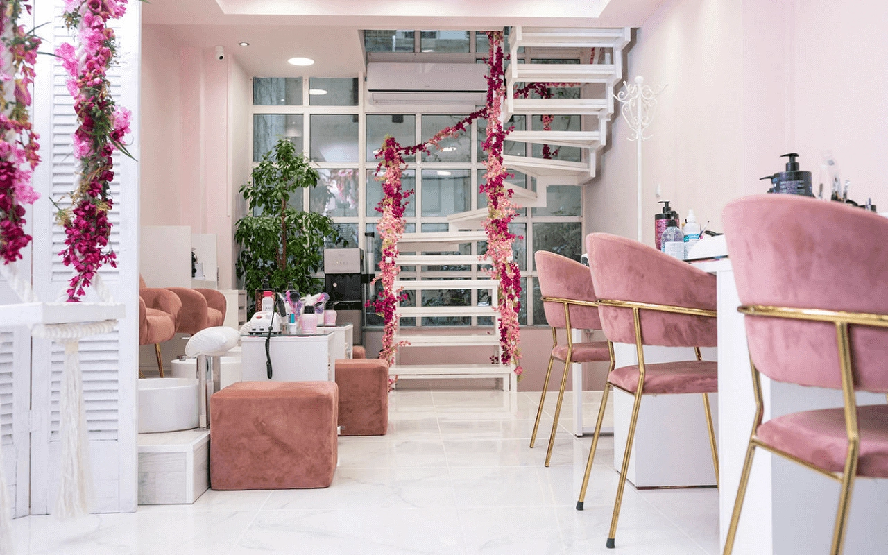 Een vrouwelijke nagelsalon met blushroze fluwelen stoelen, een witte wenteltrap omwikkeld met roze kunstbloemen, luikachtige scheidingswanden, en goudkleurige manicurestations op een marmereffect-vloer.