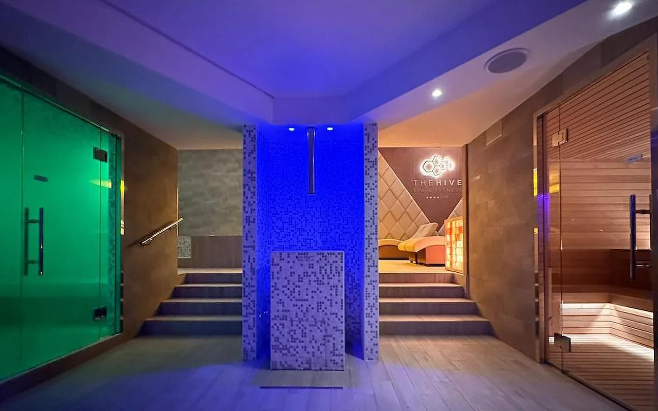 De thermische zone van een luxe spa met dramatische blauwe en groene sfeerverllichting. De ruimte heeft een houten Finse sauna, een stoombad-ingang en een centraal mozaïeken element met treden naar relaxzones.