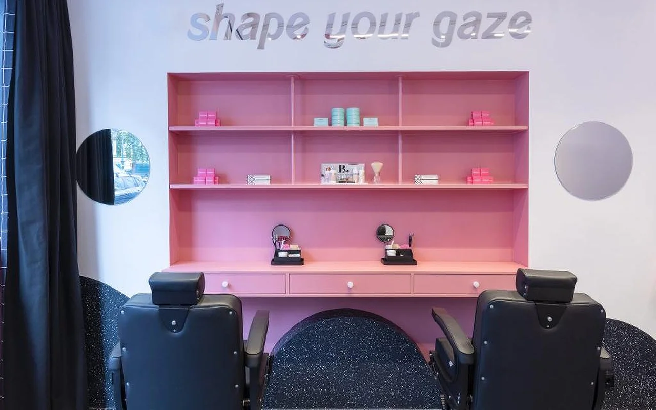 Een gespecialiseerde wenkbrauw- en wimperstudio met een opvallend roze wandrek vol beautyproducten, zilveren “shape your gaze”-letters op de muur, en zwartleren behandelstoelen op een donkere gespikkelde vloer.