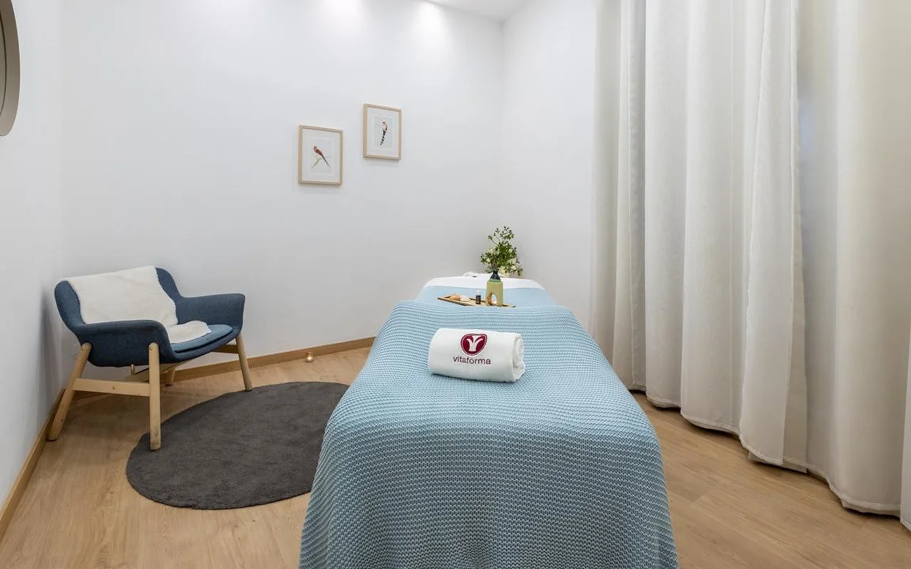 Een schone, minimalistische sparuimte met witte muren en licht houten vloeren. Een behandeltafel is netjes opgemaakt met een lichtblauwe gebreide deken en een opgerolde witte handdoek met het 'Vitaforma'-logo. In de hoek staat een stijlvolle blauwe fauteuil op een klein rond grijs kleed, en lange witte gordijnen hangen naast het raam.
