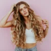 Studiofoto van een vrouw met heel lang, golvend, door de zon gekust balayage-haar, gekleed in een lichtblauwe jeans en een wit kort gebreid topje
